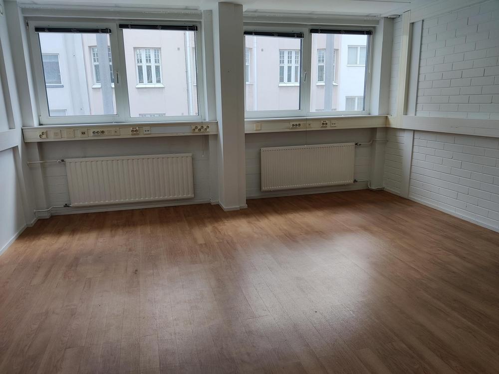 Kuva tyhjästä toimitilasta neljällä ikkunalla. Huoneen seinät ovat valkoiset ja lattia on puinen laminaatti.