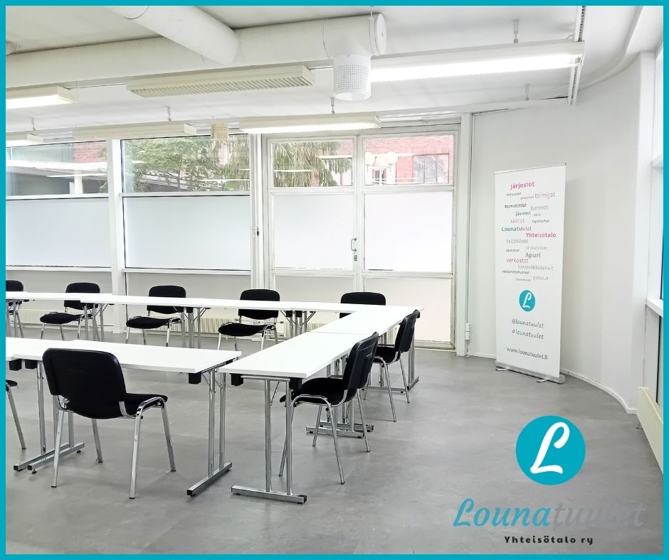 Valokuva toimitilasta, jossa valkoiset seinät, harmaa lattia, suuret ikkunat, ja keskellä huonetta pöytiä ja tuoleja ympyrämuodostelmassa.